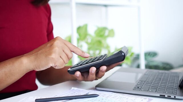 close-up of a woman  using a calculator to manage personal finances, debts, and bills with a laptop and documents on a white desk, focusing on financial planning