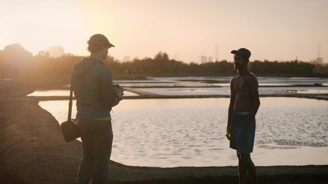 A male photographer is clicking a picture using a camera of a young daily wage worker and shakes his hand, during a golden hour in the salt pan flats filled with seawater and sunset in the background.