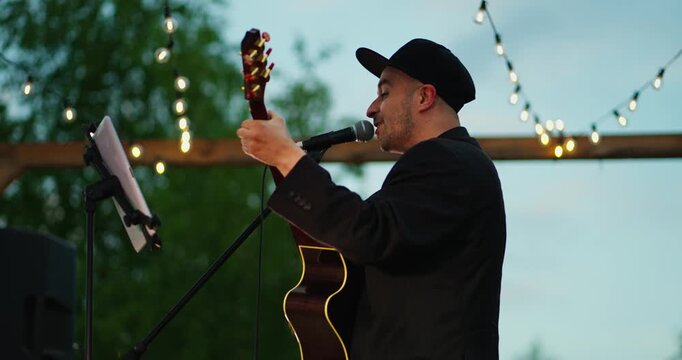 Male Acoustic Guitarist Singing Under Warm String Lights at Dusk