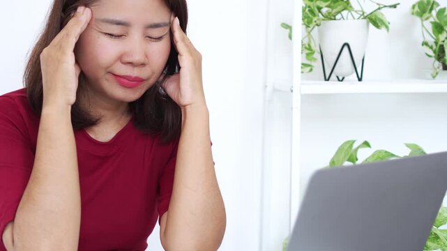 Asian woman in a red shirt touching her temples with closed eyes, feeling stressed, tired, or having a headache while working on a laptop at a bright home office desk, representing burnout