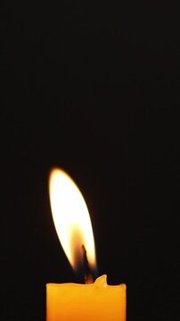 Close-up of a burning yellow candle flame glowing brightly in the dark, moving gently before being blown out with smoke rising against a black background.