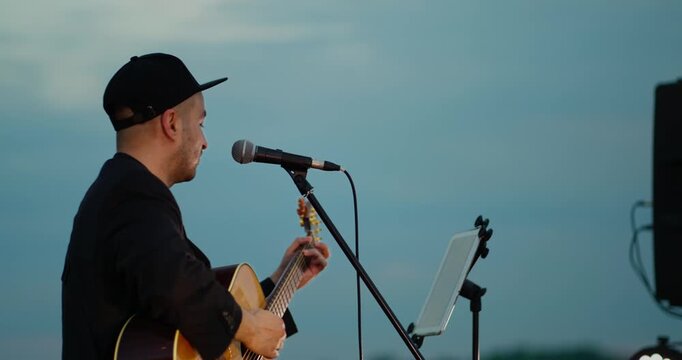 Male acoustic guitarist singing in profile on outdoor dusk stage