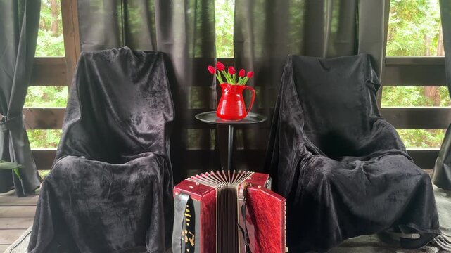 Dusty Accordion and Tulips: An old accordion rests on a rustic wooden floor, flanked by two chairs draped in dark fabric, with a vibrant red pitcher of tulips on a small table.