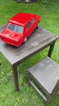 Vintage Toy Car on Table: A classic red toy car, resembling a vintage sedan, sits on a weathered wooden table outdoors, set against a backdrop of lush green grass.