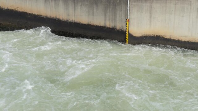 Torrent of water flowing through concrete canal, past yellow and black depth gauge, wide angle