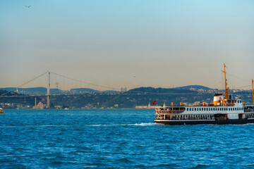 Fototapeta premium Traditional Passenger Ferry Boat Cruising on the Bosphorus Strait with the Iconic Bosphorus Bridge and City Skyline of Istanbul, Turkey under a Clear Blue Sky at Daytime.