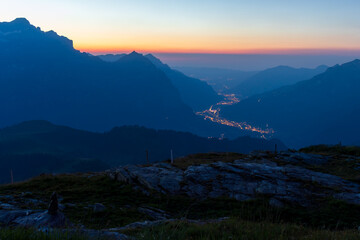 Beautiful view of mountain peaks during twilight, with a winding valley of glowing city lights nestled between slopes under a vibrant sunrise sky. © Adrian