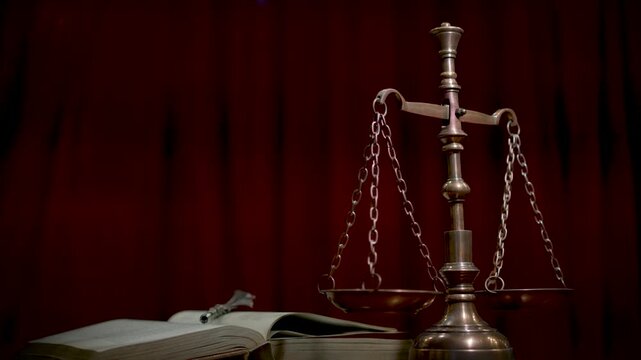 A close-up of a brass scale of justice with balanced pans on a wooden surface against a dark red curtain in a classic legal atmosphere.
