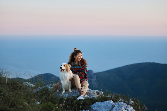 A woman relaxes with her Border Collie on a rocky mountain viewpoint above the sea. Natural scenery creates outdoor lifestyle and adventure atmosphere.