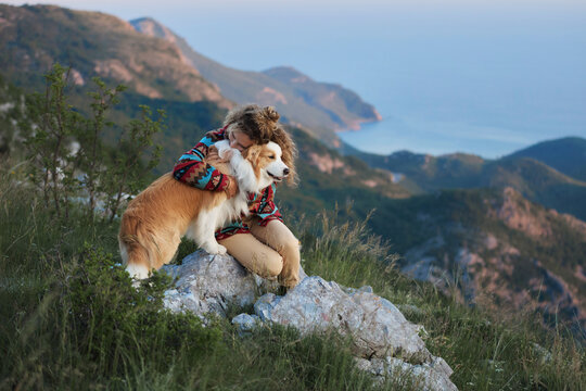 A woman embraces her Border Collie while sitting on rocky terrain above mountain scenery. Warm atmosphere creates outdoor travel inspiration.