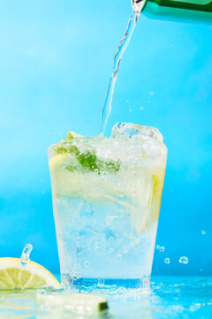 Pouring refreshing carbonated drink into glass with lime and ice on blue background
