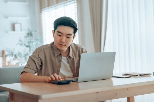 Happy young Asian man working on laptop in living room. Freelance worker male sitting at desk writing notes while watching webinar, studying online