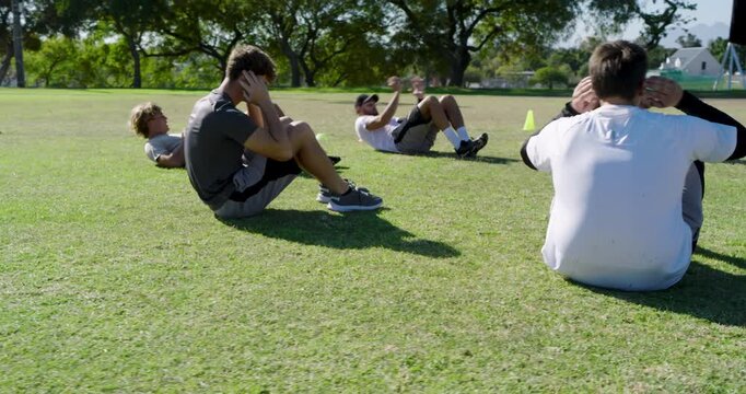 Performing crunches diverse male group on grass field in park, yellow training cones