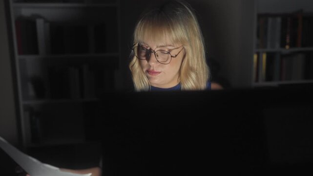 Woman removing glasses and rubbing neck in dim studio with computer glow and bookshelf behind; late night fatigue.