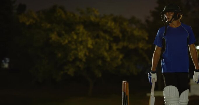 Teenage white batsman entering crease by floodlights, pausing sizing field with partner copy space