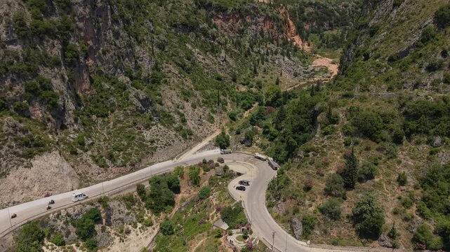 Aerial view of a winding mountain road with vehicles navigating a sharp hairpin turn through a lush green canyon and rocky cliffs in Dhermi, Vlore County, Albania.