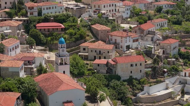 Aerial view of the village of Dhermi featuring traditional red-tiled roofs and a white church with a blue dome nestled among green trees in Dhermi, Vlore County, Albania.