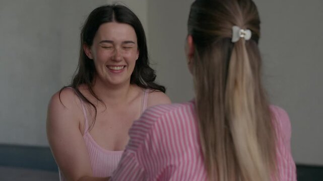 Two women embrace warmly, sharing smiles and comfort. One wears a pink striped shirt, the other a soft pink top. Their laughter and closeness suggest friendship or familial bond