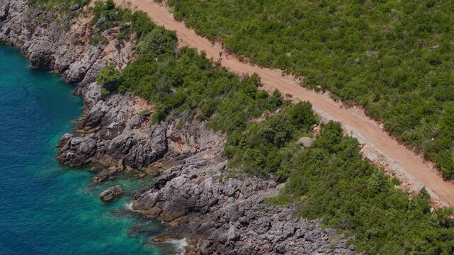 Aerial view of a dirt road winding along the rugged rocky coastline and turquoise waters near Aquarium Beach, Jala, Vlore County, Albania.