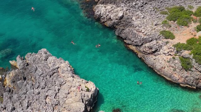 Aerial view of people swimming in the turquoise waters of Aquarium Beach surrounded by rocky cliffs and green shrubs in Jala, Vlore County, Albania.