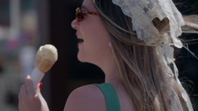 Woman in lace headpiece enjoys ice cream outdoors. Sunglasses frame her calm, contented expression. Soft breeze lifts her hair gently. Sunlit streets suggest a pleasant day. Ideal for travel, festival