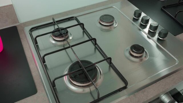 Hand cleans stove burner with green cleaning sponge. Stove has four burners with black grates. Person uses gentle motion for thorough cleaning. Surface gleams clean after wiping
