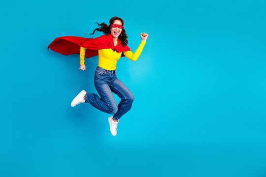 Young woman superhero jumps in the air wearing a red cape and yellow shirt against a blue background