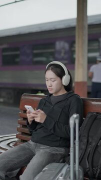 Traveler sitting on a bench using smartphone with headphones at a train station, ideal for travel lifestyle, waiting, mobile app, music and railway journey concepts.