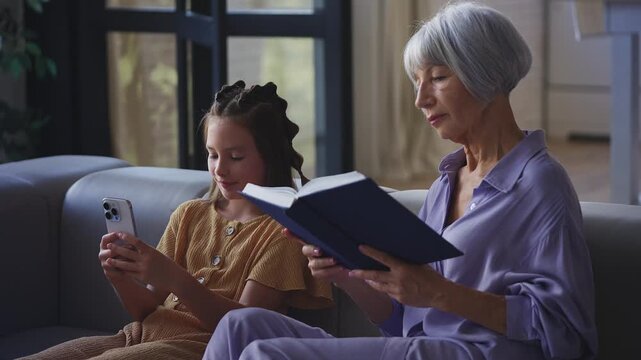 Grandmother reading her favorite book while her granddaughter plays on a phone