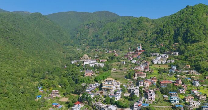 Aerial slow-orbit shot of houses and a Guru Rinpoche statue nestled among the green hills and vibrant farming fields of Pharping, Nepal, under a clear blue sky.