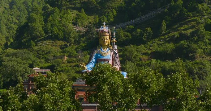 Tilt up reveal shot from green leaves and branches uncovers the grand Guru Rinpoche statue in Pharping Nepal against a lush hillside in a bright sunny day.