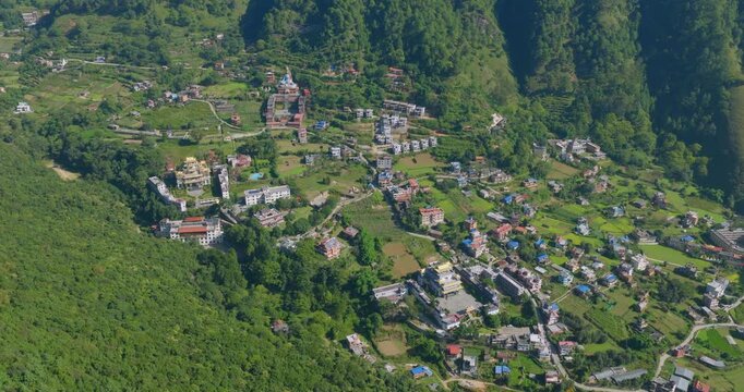 Aerial orbit shot captures a scenic view of tiny houses nestled among the green hills and farming fields of Pharping Nepal under a clear blue sky.
