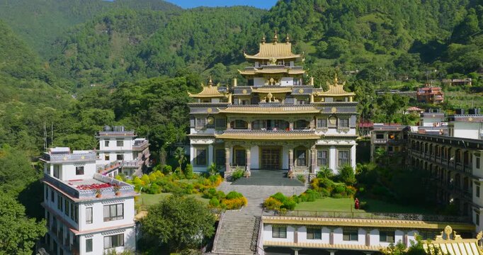 Aerial slow push in shot approaches the golden Rigon Tashi Choeling Monastery in Pharping Nepal, moving toward the backside against lush green hills on a bright sunny day.
