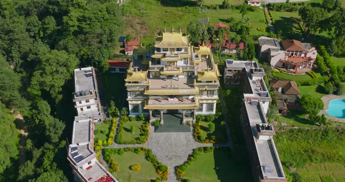 Aerial tilt down shot of the golden architecture of Rigon Tashi Choeling Monastery in Pharping Nepal, surrounded by lush green hills and houses in the background