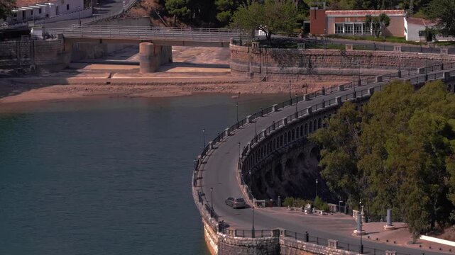 Drone footage orbiting forward around El Chorro's sweeping dam road, tracking a lone car rounding the curved stone parapet above jade water, sandy shore, and a cylindrical intake tower.