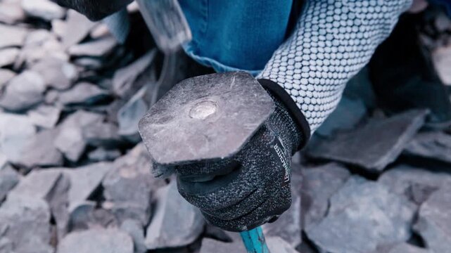 A slow-motion, macro close-up shot of a geologist's gloved hands striking a flat-headed chisel with a geological hammer. Tiny fragments and dust erupt as the tool impacts a layered slab of grey slate,