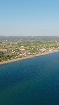 Slow vertical panoramic drone pan of Kourouta beach, Greece. Beach cafe bar umbrellas, bathers on shore. Clear sunny summer day