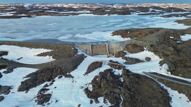 Mid-range drone shot showing a reinforced concrete dam structure and water control spillway surrounded by snow-patched tundra.