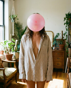Anonymit&auml;t und Identit&auml;t: Kreatives Portr&auml;t einer Frau mit rosa Ballon vor dem Gesicht im hellen Zimmer.