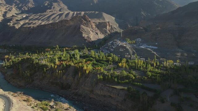 The Indus River curves around Takmachik village near Leh in Ladakh, India, with green poplar groves contrasting against barren mountain slopes. Smooth aerial tracking shot reveals the river valley.