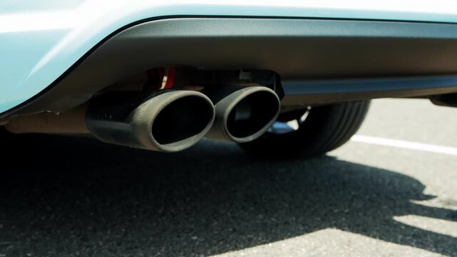 Detail of two dark exhaust pipes, rear of light colored car. Pipes integrated into black bumper element, showing metallic texture. Vehicle parked asphalt under bright sunlight, suggesting outdoor sett