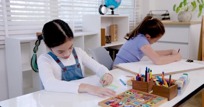 Asian girl in white shirt and caucasian classmate focusing on drawing colorful art in notebook, elementary students studying creativity at school desk with pencils and stationery, teamwork learning