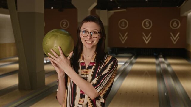 Portrait of Caucasian young zoomer woman with glasses holding bowling ball and smiling at camera in bowling alley. Perfect for promoting indoor sports, active lifestyle, and weekend leisure