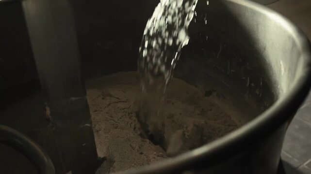 Close up view of baker pouring cold water to dry flour in large metal bowl of commercial spiral dough mixer. Mixing ingredients for fresh bread dough, essential step of baking process. Baker pours ice