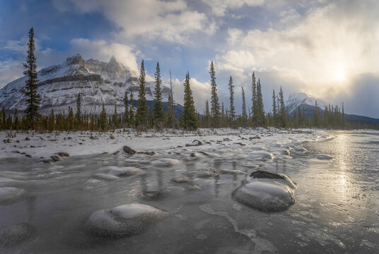 View of a frozen lake with ice bubbles and snow-capped mountains under a golden sun along the Icefields Parkway in Jasper, Alberta, Canada.