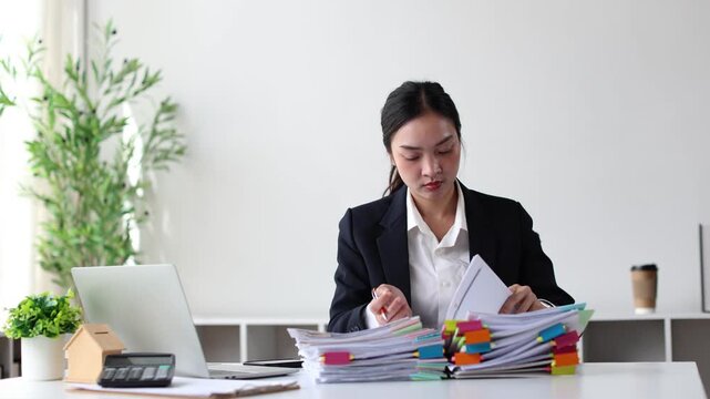 Asian businesswoman sorting through a large stack of documents and paperwork at her office desk, managing financial reports and signing important contracts for a new project