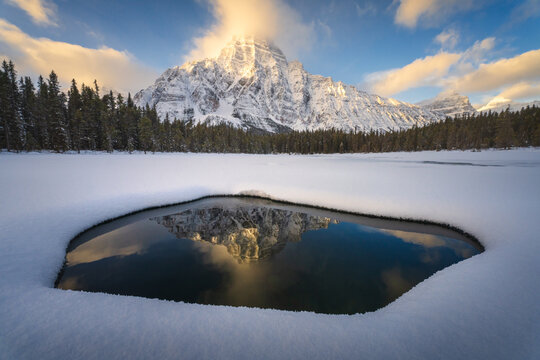 View of a snow-capped mountain peak reflected in a clear pool of water amidst a vast snowy field and evergreen forest along the Icefields Parkway in Jasper, Alberta, Canada.