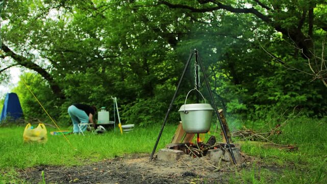 Outdoor cooking activity in summer forest, person tending camp, pot hangs over open fire on tripod, steam rises from pot, lush green trees and grass provide natural backdrop, camping lifestyle food pr