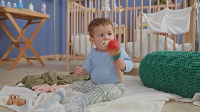 Boy sitting on blanket holding a wooden maraca, pacifier in mouth and hand gripping toy, inside a building nursery scene; gentle curiosity.