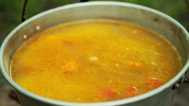 Close-up view of large metal pot on a hook, containing bubbling soup or stew. Warm broth with visible pieces of sausage and diced carrots, cooking outdoors. Rustic meal preparation during summer outdo